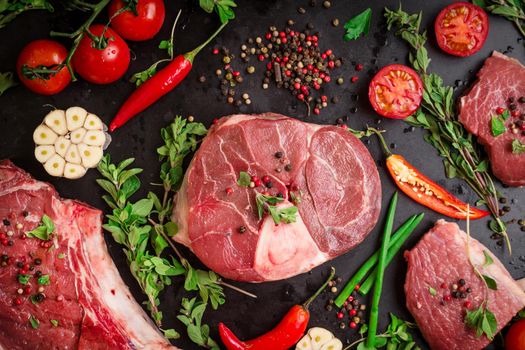 Different types of steaks set. Assorted raw meat on a black chalk board background. Rib eye steak on the bone, veal shank (ossobuco), fillet with cherry tomatoes, hot pepper and herbs.