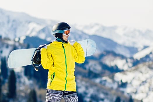 Young woman snowboarder with snowboard on her shoulders looks at the at the ski slope, copy space