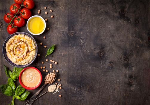 Bowl with hummus, chickpea, tahini, olive oil, sesame seeds, cherry tomatoes and herbs on dark rustic wooden background. Space for text. Food frame. Middle eastern cuisine. Top view. Hummus background