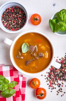 Delicious tomato soup with meat on a white rustic wooden table with fresh cherry tomatoes, basil leaves, dry pepper and red gingham kitchen towel. Ingredients for soup. Top view