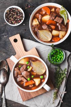 Meat stew with beef, potato, carrot, pepper, spices, green peas. Slow cooked meat stew, bowl, wooden background. Hot autumn/winter dish. Closeup. Top view. Comfort food. Homemade soup/ragout/casserole