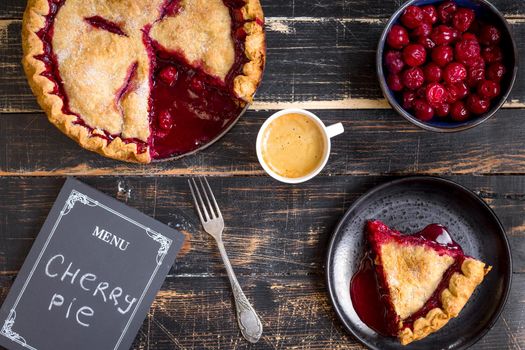 Homemade sliced cherry pie with flaky crust, cup of coffee, bowl with cherries and menu chalkboard on the black wooden table. Top view