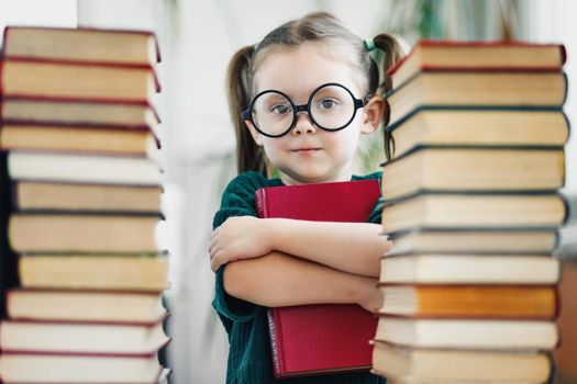 Preschool age girl in big glasses holding red book among book bunches.