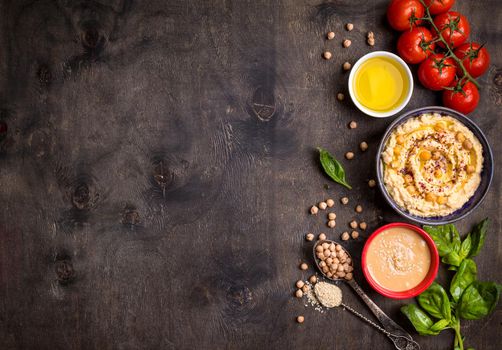 Bowl with hummus, chickpea, tahini, olive oil, sesame seeds, cherry tomatoes and herbs on dark rustic wooden background. Space for text. Food frame. Middle eastern cuisine. Top view. Hummus background