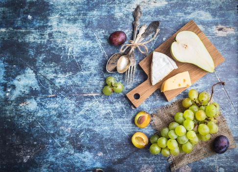 Fresh ripe pear, assorted cheeses platter, grapes, plums in bowl, blue wooden rustic background. Space for text. Top view. Fruits, Camembert, Emmental cheeses. Delicacy food. Snack/appetizer