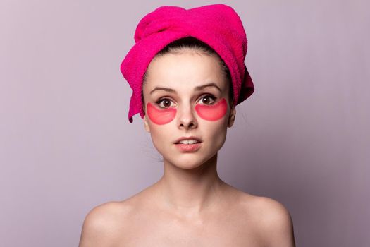 brunette woman with cosmetic pink patches under her eyes in a pink towel on her head, gray studio background, close-up portrait. High quality photo