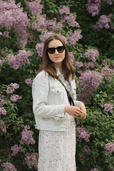 A woman is staying near the lilac bush in the village green. A lady in black sunglasses is posing in the park.