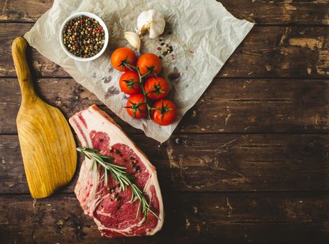 Raw marbled meat steak, pepper, herbs, tomato, old wooden background. Space for text. Beef Rib eye steak ready for cooking. Top view. Copy space. Ingredients, meat roasting. Ribeye meat steak. Closeup