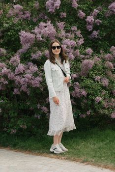 A woman with long hair is staying near the lilac bush in the village green. A lady in black sunglasses and white dress is posing in the woodland.