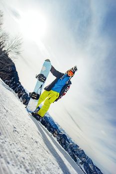 Bearded happy snowboarder in a ski mask with goggles and a fur big mohawk hat on a background of a sky and winter snowy mountains