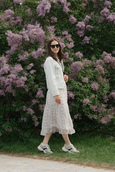 A woman with long hair is staying near the lilac bush in the village green. A lady in black sunglasses and white dress is posing in the park.