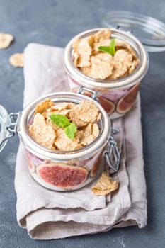 Homemade healthy yoghurt in glass pot with cereals, figs, mint on rustic concrete background. Healthy morning breakfast. Freshly made yoghurt with oat granola and fruit. Yoghurt in jar with muesli