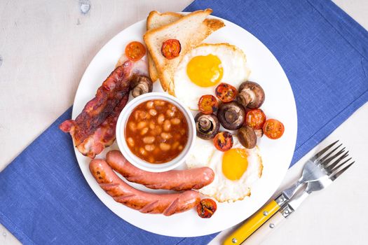 Full english breakfast with fried eggs, tomatoes, sausages, bacon, mushrooms, toasts and beans. Breakfast on a white plate with forks and blue napkin on a white wooden table. Top view