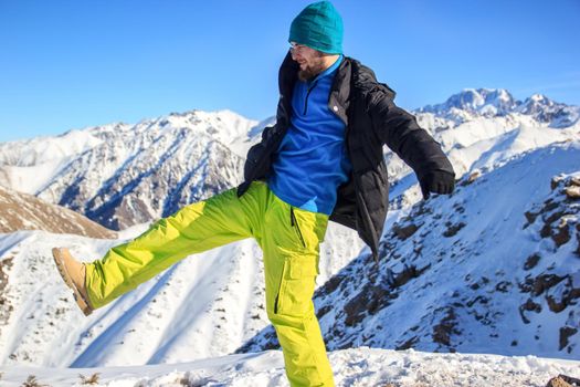 A bearded man in colorful winter clothes fooling around on a mountain top. The joy of conquering the top, adventure and travel.