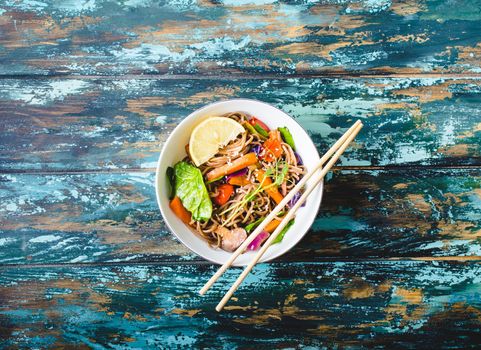 Asian noodles with vegetables, bowl, rustic wooden old background. From above. Top view. Soba noodles, vegetables, mushrooms, chopsticks. Vegetarian/Vegan noodles. Asian style dinner with noodles