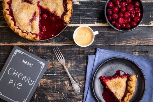 Homemade sliced cherry pie with flaky crust, cup of coffee, bowl with cherries and menu chalkboard on the black wooden table. Top view