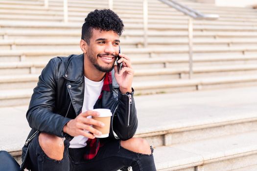 happy young latin student sitting on a staircase smiles talking on the phone with a coffee in his hand, concept of technology and urban lifestyle