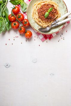 Italian pasta bolognese. Spaghetti with meat and tomato sauce in a plate with Italian tablecloth on a wooden white background. With fresh cherry tomatoes, basil. Food frame. Space for text. Top view