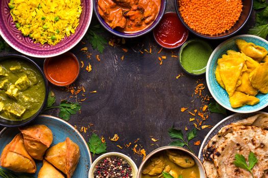 Assorted indian food on dark wooden background. Dishes of indian cuisine. Curry, butter chicken, rice, lentils, paneer, samosa, naan, chutney, spices. Space for text. Bowls and plates with indian food