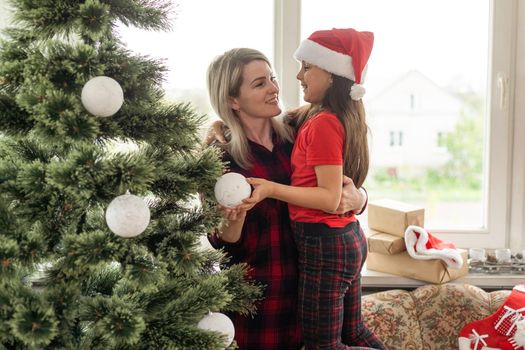 Attractive mother and daughter wearing winter outfits and decorating Christmas tree at home with joy, preparing to celebrate Christmas festival. Concept for family Christmas party