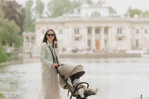 A mother is posing with her baby in the stroller on the territory of the palace. A mom in sunglasses with a baby in the carriage with classical architecture in the background in the park.