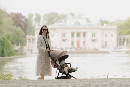 A mother in full growth is posing with her baby in the stroller on the territory of the palace. Mom in sunglasses with a baby in the carriage with classical architecture in the background in the park