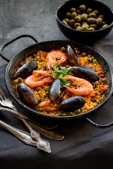 Paella in black pan with rice, shrimps, mussels, squid and meat, bowl with olives and vintage cutlery. Seafood paella, traditional spanish dish. Paella on rustic black wooden table. Selective focus