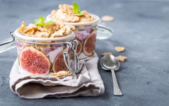 Homemade healthy yoghurt in glass pot with cereals, figs, mint on rustic concrete background. Healthy morning breakfast. Freshly made yoghurt with oat granola and fruit. Yoghurt in jar with muesli