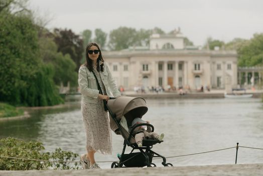 A mother is posing with her baby in the stroller on the territory of the palace. A mom with a baby in the carriage with classical architecture in the background in the park.