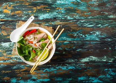 Traditional Vietnamese soup Pho bo with herbs, meat, rice noodles, broth. Pho bo in bowl with chopsticks, spoon. Space for text. Top view. Asian soup Pho bo on wooden table background. Vietnamese soup