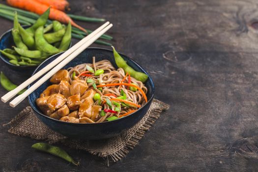 Asian noodles with chicken, vegetables in bowl, rustic wooden background. Space for text. Soba noodles, teriyaki chicken, edamame, chopsticks. Closeup. Asian style dinner. Chinese/Japanese noodles