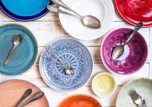 Different vintage multicolored empty plates and bowls on a white old wooden table. With antique spoons and forks. Table setting. Shabby chic/retro style. Top view. Rustic kitchen. Set of plates