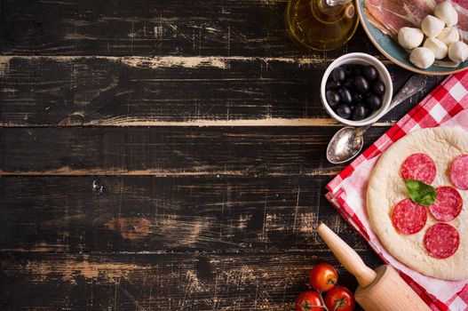 Pizza making background. Ingredients for making pizza. Space for text. Pizza dough, flour, cheese, mozzarella, tomatoes, basil, pepperoni, olives and rolling pin over black wooden background. Top view