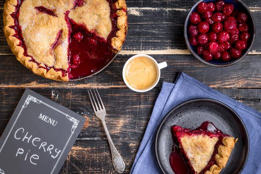 Homemade sliced cherry pie with flaky crust, cup of coffee, bowl with cherries and menu chalkboard on the black wooden table. Top view