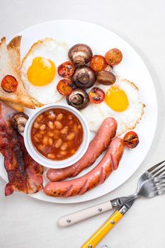 Full english breakfast with fried eggs, tomatoes, sausages, bacon, mushrooms, toasts and beans. Breakfast on a white plate with forks on the white wooden table. Top view