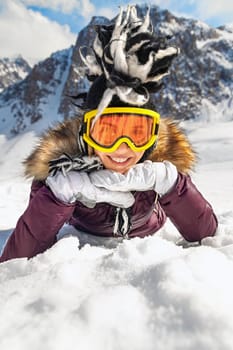 Portrait of playful hipster woman lying on snowy at mountain or hill top in sunny day with blue sky and big rock view in background. Winter holidays, tourism, travel concept.