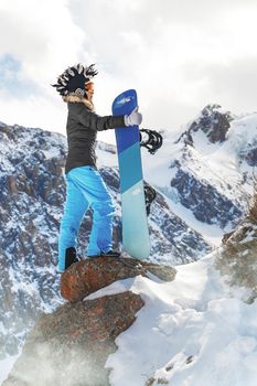 Portrait of active female snowboarder in funny hat standing on top of the mountain background. Copy space, vertical