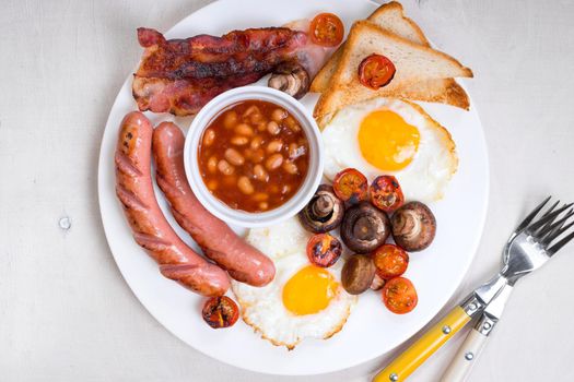 Full english breakfast with fried eggs, tomatoes, sausages, bacon, mushrooms, toasts and beans. Breakfast on a white plate with forks on the white wooden table. Top view