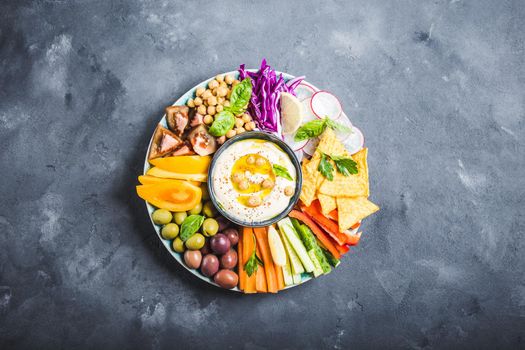 Hummus platter with assorted snacks. Hummus in bowl, vegetables sticks, chickpeas, olives, pita chips. Plate with Middle Eastern/Mediterranean meze. Party/finger food. Top view. Vegetables, hummus dip