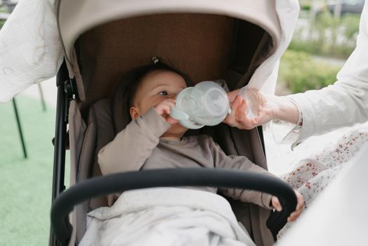 An infant baby in the stroller is drinking from her bottle which is holding her mother.