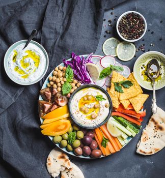 Meze platter with hummus, yoghurt dip, assorted snacks. Hummus in bowl, vegetables sticks, chickpeas, olives, pita, chips. Plate with Middle Eastern/Mediterranean meze. Party/finger food. Top view