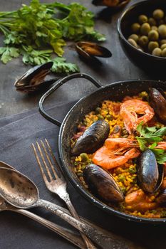 Paella in black pan with rice, shrimps, mussels, squid and meat, bowl with olives and vintage cutlery. Seafood paella, traditional spanish dish. Paella on rustic black wooden table. Selective focus