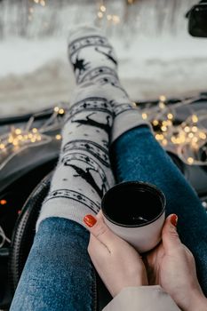 Woman legs with socks hand holding thermos cup of coffee or tea in winter car. Christmas socks. Garland defocused lights. Winter travel.