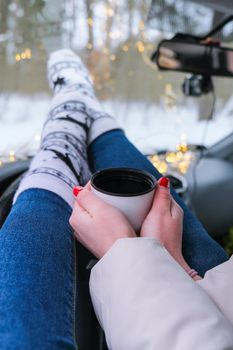 Woman legs with socks hand holding thermos cup of coffee or tea in winter car. Christmas socks. Garland defocused lights. Winter travel.