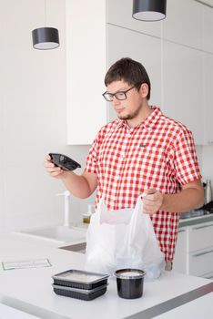 Delivery food, products to home. Shopping and healthy food concept. Young man in red plaid shirt holding a disposable plastic bag with food delivery at the modern kitchen