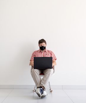 Social distancing. E-learning. Young man in black protective mask using laptop, sitting at the chair. Copy space