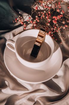 White cup of coffee with piece of chocolate inside. Morning aesthetics vibes. Breakfast. Pink gypsophila flowers. Hard shadows.