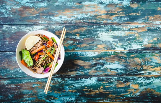 Asian noodles with vegetables, bowl, rustic wooden old background. Space for text. Top view. Soba noodles, vegetables, mushrooms, chopsticks. Vegetarian/Vegan noodles. Asian style dinner with noodles