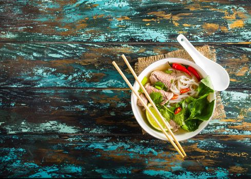 Traditional Vietnamese soup Pho bo with herbs, meat, rice noodles, broth. Pho bo in bowl with chopsticks, spoon. Space for text. Top view. Asian soup Pho bo on wooden table background. Vietnamese soup