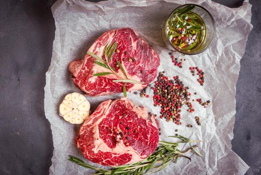 Raw juicy steaks with seasonings on a baking paper ready for roasting on rustic concrete background. Fresh marbled meat steaks with herbs, garlic, olive oil, colorful whole pepper, rock salt. Top view